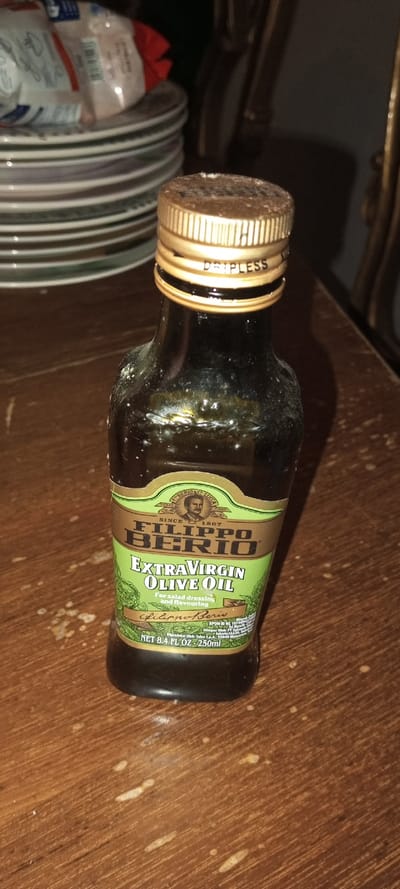 An olive oil bottle stands prominently on a wooden surface. The bottle, labeled "Filippo Berio Extra Virgin Olive Oil," features a green and gold label. The bottle's cap is gold, with "Dripless" printed on it. To the left of the bottle, a stack of plates and what appears to be snack bags is visible. The wooden surface has small, scattered droplets. The photo's lighting is dim, casting shadows on the scene.