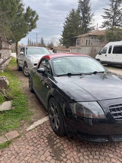The scene is set on a street in Bryastovo, Bulgaria, under an overcast sky. In the foreground, a black Audi TT convertible with a red hardtop is parked on a cobblestone surface, angled slightly towards the right, showcasing its front and left side. The vehicle's headlights appear darkened or tinted. Behind the Audi TT and to its left, a silver Porsche Cayenne SUV is parked on a dirt and grass verge alongside a low, dry-stacked stone wall. Further in the background on the right, a white Ford Transit-style passenger van is partially visible behind a concrete wall. An older residential building with a tiled roof and visible chimneys is situated in the mid-background, surrounded by various evergreen and bare deciduous trees. A utility pole stands on the left side of the background. No individuals are present in the scene, and all vehicles are stationary.