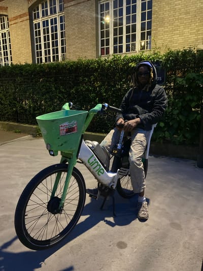 A man with dreadlocks, wearing a blue head covering and a dark jacket, sits on a Lime brand e-bike on a paved surface. He is holding the handlebars and looking towards the camera. The e-bike has a green basket in front. In the background are a brick building with large windows, some bushes, and a sign reading "Arrêts". The location is likely Le Pré-Saint-Gervais, France.
