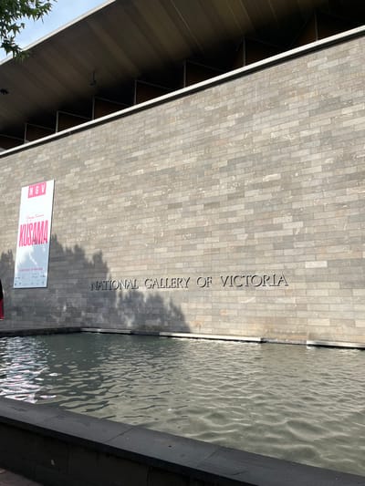The image captures the exterior of the National Gallery of Victoria building in Southbank, Australia, on a bright, sunny day. The main subject is the building's facade, constructed from light grey, rectangular stone or brick tiles. The words "NATIONAL GALLERY OF VICTORIA" are carved or embedded in dark lettering along this textured wall.
Adjacent to the building is a long, narrow water feature, likely a moat or decorative pool, with a gently rippled surface reflecting the sunlight. Shadows of nearby trees are cast across the building's wall and the water, indicating the time of day.
An angled roof structure, composed of dark panels with visible supports, is seen on the left side of the building. A white banner with pink highlights is affixed to the facade, displaying "NGV" and the name "KUSAMA," referring to artist Yayoi Kusama. The banner also indicates an exhibition running from "15 DEC 2023 - 21 MAY 2024," along with various logos and additional small text. Green foliage from a tree is visible in the bottom left, and a sliver of blue sky can be seen. No people are present in the scene.