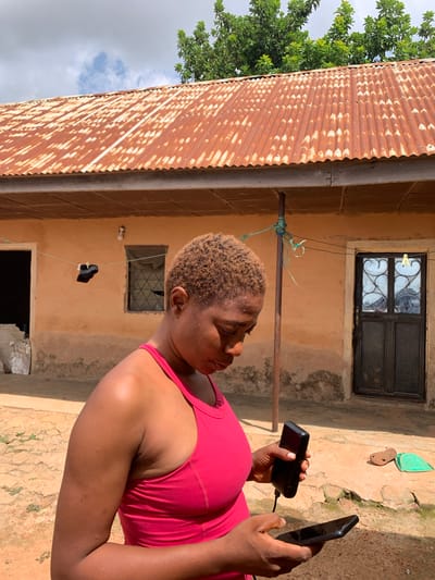 A woman with short, blond hair is holding a black smartphone and a black power bank. She is wearing a pink tank top and is standing in what appears to be an outdoor courtyard in front of a light brown, stucco-walled building. A corrugated metal roof, partially rusted, is visible to the left, with green foliage and a cloudy sky above. The building has a dark-colored metal-framed window with decorative bars above a smaller, open window. A clothesline with a single piece of dark fabric hangs in front of the building. The ground is dirt and cracked concrete. The lighting suggests it is daytime. The scene is likely in Russo, Nigeria.