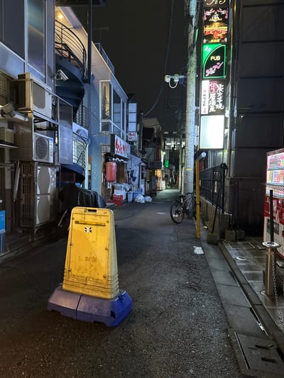 The image shows a narrow, dimly lit alleyway at night in Adachi, Japan. The alley is flanked by multi-story buildings with various signs and air conditioning units visible. In the foreground, a bright yellow traffic cone with a blue base stands in the middle of the asphalt road, which appears wet. To the left, a metal staircase spirals up the side of a building. On the right side of the alley, a black bicycle is parked next to a pole with a yellow vertical marker. Further down the alley, a vending machine is visible with a chain barrier in front of it. Several trash bags are piled up near the buildings on the left side. The overall impression is a quiet, urban backstreet at night.