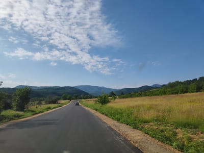 A paved road curves through a rural landscape under a bright blue sky dotted with white clouds. The road is dark asphalt with a gravel shoulder and some patchy green grass and dry weeds along its edge. On the left side of the road, there are trees and rolling green hills in the distance. On the right side, a field of tall, dry, golden grass stretches out, also with some green vegetation at its base. In the distance, a line of mountains forms the horizon. A small, dark-colored car is visible on the road, driving away from the viewer and towards the mountains. The image appears to be taken from a moving vehicle, looking forward along the road. The scene suggests a journey through a scenic, countryside area in the vicinity of Lesidren, Bulgaria.