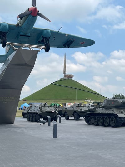 This media file captures an outdoor exhibition of military hardware, likely a museum or memorial. The foreground features a light blue aircraft with a red star emblem, suspended above a dark grey structure. The aircraft has a prominent propeller and appears to be a Soviet-era fighter, possibly an Ilyushin Il-2, as suggested by the text on the structure.
Behind the aircraft, a grassy, sloped mound forms the backdrop, culminating in a sharp, metallic spire-like monument. Along the base of the mound, a line of grey military vehicles, including tanks and what appear to be armored personnel carriers, are displayed. The vehicles have red star emblems visible on their hulls. Small tents in various colors are visible behind the vehicles, suggesting a public event or a visitor area.
The sky is partly cloudy with a bright, daytime light, indicating it is likely midday. The ground is paved with light grey square tiles. The overall impression is one of commemoration and historical display.
Visible text on the grey structure reads: "ЛЕГЕНДАРНЫЙ ШТУРМОВИК ИЛ-2". This translates to "LEGENDARY ATTACK AIRCRAFT IL-2".
The setting is consistent with the Ozyaritska-Slabadski rural council in Belarus, an area known for its historical significance, particularly related to World War II. The presence of both an aircraft and tanks suggests a focus on ground and air warfare. No people are clearly visible in the image, though some may be present out of frame or too small to discern.