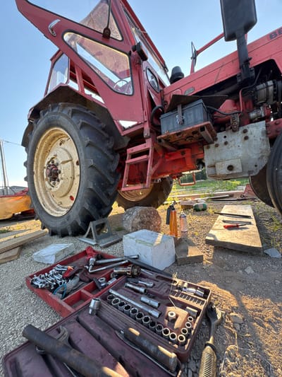 A red tractor is parked on gravel, with its cab door open. Tools, including a socket wrench set in a red case, are laid out on the ground next to the tractor. A car battery is visible under the hood. The scene appears to be outdoors in daylight, possibly late afternoon, with the sun casting long shadows. The location is Rezekne, Latvia. There is no visible text in the image.
