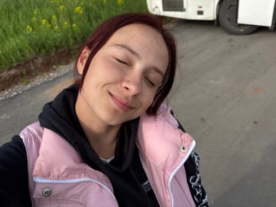 A woman with reddish-brown hair and closed eyes is centrally framed. She is wearing a black hoodie with the hood up, and a pink puffer jacket. The background consists of pavement and grass with small yellow flowers. A tire and a white object, possibly a vehicle, are visible in the lower left corner. The photograph is taken from an overhead angle. There is no verifiable information related to the location of Krilovo, Russia in the given image.