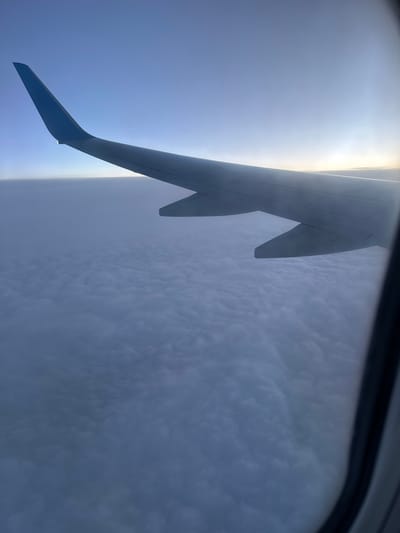 The image shows a view from an airplane window. Part of the airplane wing is visible, with blue and grey coloration. Beyond the wing, there's a dense cloud formation. The presence of the wing suggests the image was taken during a flight. The location (Kitsjanovo, Russia) cannot be confirmed from the image's visual information. There is no visible indication of any people, objects, actions, or interactions. The environment consists of the sky, clouds, and the airplane wing.