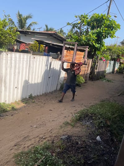 A man is carrying a large, pink plastic chair over his head and shoulders, on a dusty dirt road in a semi-urban area. He is wearing a dark green polo shirt and blue shorts. The chair is wrapped in a red and white cloth at the top. To his left is a corrugated metal fence, and behind that, buildings with corrugated roofs and palm trees are visible. To his right, there is a wooden fence and more greenery. The sky is clear and blue. The scene appears to be in Moratuwa, Sri Lanka, a coastal city known for its furniture manufacturing.