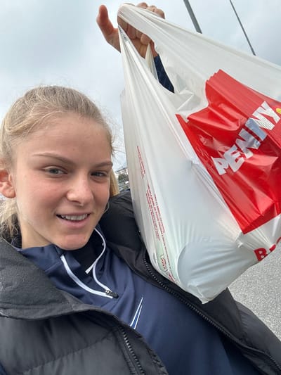 A young adult female is depicted in a selfie, smiling directly at the camera. She has blonde hair pulled back and is wearing a navy blue quarter-zip top with a white Nike swoosh logo on the chest, beneath an unzipped black puffer jacket. Her right hand, positioned at the top center of the frame, firmly grips the handles of two white plastic shopping bags. The front bag prominently displays red branding with the text "PENNY," indicating a purchase from a Penny Market supermarket. The background is out of focus, showing a predominantly overcast sky and indistinct outdoor structures. The scene is consistent with an individual having completed shopping, likely in a street or parking lot environment within the city of Borgo San Dalmazzo, Italy.