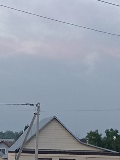 The image shows a view of houses and trees under a cloudy sky, with utility wires crossing the frame. In the foreground, a utility pole with wires attached is visible, leaning slightly. Behind the pole, the roofline of a light-colored, gable-roofed house is prominent, with horizontal siding. To the left of this house, another smaller house with a grey roof is partially visible. Further back, a line of green trees forms a backdrop. The sky is overcast with some lighter areas, suggesting diffused light. The scene appears to be in a rural or suburban setting.