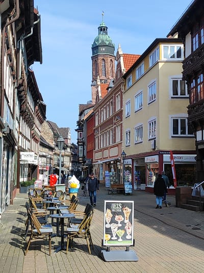 The image depicts a bustling pedestrian street in the city of Einbeck, Germany, with the Marktkirche St. Jacobi's green-domed tower prominent in the background under a clear blue sky. On the left side of the cobbled street, a row of traditional half-timbered buildings with ground-floor shops features outdoor cafe seating, including six dark circular tables surrounded by woven chairs. A large, stylized inflatable ice cream cone, yellow and white, stands near the cafe tables. Further down the street, more historic buildings line the pathway. On the right, a multi-story building with a pale yellow facade and numerous white-framed windows is visible, along with storefronts displaying "Porzellan" and "HINNERT."
Several individuals are present: a man in a dark jacket and jeans walks towards the viewer on the right side of the street, near a bicycle. Further back, a couple walks away from the viewer, one person in a dark jacket and blue jeans, accompanied by a small, light-colored dog. Additional indistinct figures are visible in the mid-ground. In the immediate foreground, on the right, a gray sandwich board sign advertises "EIS BECHER" (ice cream sundaes) with photographic examples. Streetlights, benches, and various shop displays are also distributed along the street.