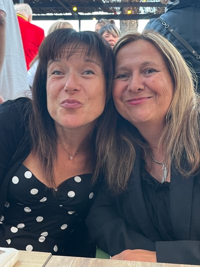 Two women are posing for a selfie in an outdoor setting, likely a restaurant or event space. The woman on the left has shoulder-length dark brown hair with bangs and is wearing a black and white polka dot top with a black cardigan. She has a delicate necklace with a small pendant. The woman on the right has shoulder-length blonde hair and is wearing a black jacket over a black blouse, also with a necklace, this one a more elaborate silver chain with dangling gems. Both women are smiling and looking at the camera. In the background, other people are visible, including a person in a red jacket. The environment appears to be somewhat festive, with some lighting in the background. The table in the foreground is light brown wood. Based on the context provided, this image was taken in the city of Saint-Priest-en-Jarez, France.