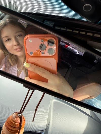 The image is a selfie taken in a car's rearview mirror. The main subject is a young woman, visible from the chest up, her face reflected in the mirror. She is holding an orange-cased smartphone in her right hand, which is positioned to take the picture. Her left hand is also visible, with a light yellow nail polish. She appears to be wearing a light-colored striped top.
The setting is the interior of a car. The rearview mirror is prominently featured, reflecting the woman, her phone, and parts of the car's interior. The background of the reflection shows the dashboard, with a textured surface and a visible air freshener hanging from the mirror. The air freshener is made of wood and has a string attached. There's also a glimpse of the car's front passenger seat and a person's legs, suggesting another occupant in the car.
The lighting suggests it is daytime, likely a sunny day given the bright reflections. The woman's expression is neutral, perhaps a little tired or contemplative, as she looks into the mirror. A small object, possibly a small fire extinguisher or a similar cylindrical item with red and white markings, is visible on the dashboard behind the phone. The image seems to be taken casually, possibly during a drive.
There is some faint text on the cylindrical object, but it is too blurry to read. The overall impression is a candid moment captured within the car. The location context of Petukhovo, Russia, does not provide specific visual cues within the image itself, but it grounds the setting geographically.