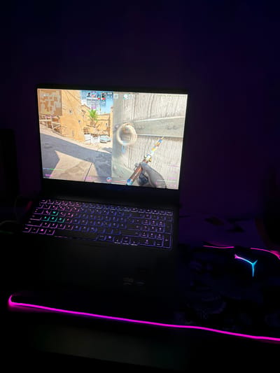 In a dimly lit room, a gaming laptop is open, displaying a first-person shooter game, likely "Counter-Strike 2," judging by the interface elements like the minimap, player icons, and in-game currency. The laptop's keyboard is illuminated with vibrant purple and pink RGB lighting. To the right of the laptop, a gaming mouse with a distinctive blue LED accent rests on a patterned mousepad, which also features RGB lighting along its edge. The overall setting suggests a personal gaming setup. No specific location in Sankt Augustin, Germany, can be determined from the provided image.