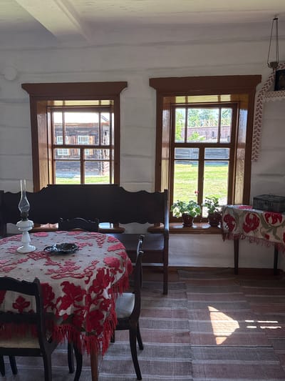 The image depicts the interior of a room in the Museum-Estate "N.V. Kopylov's Estate" in Shushenskoye, Russia. The room features rustic wooden furniture and traditional decorations. In the foreground, a round table is covered with a red and white lace tablecloth adorned with floral motifs and a fringed edge. On the table sits a white oil lamp and a small dark metal ashtray. Two wooden chairs with upholstered seats are positioned at the table. Behind the table, a dark wooden bench with a carved backrest is placed against the wall.
Two large windows with dark wooden frames are visible on the white-plastered wooden walls. The window on the left looks out onto a courtyard with traditional wooden buildings. The window on the right offers a view of a green lawn and more wooden structures, with potted plants on the windowsill. A small wooden table with a red and white patterned cloth, and a dark chest are placed to the right of the right window. The floor is covered with striped rugs, and sunlight streams through the window, casting geometric shadows. The walls are constructed from rough-hewn logs, and the ceiling has exposed wooden beams.