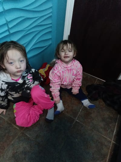 Two young girls are seated on a tiled floor near a textured turquoise wall. One wears a black and white hoodie with pink pants and grey socks. Her expression is solemn. The other girl wears a pink tie-dye hoodie and dark pants with mismatched socks. She is smiling slightly. A red item is partially visible behind them, near a dark door. A dark item lies on the floor next to the door.