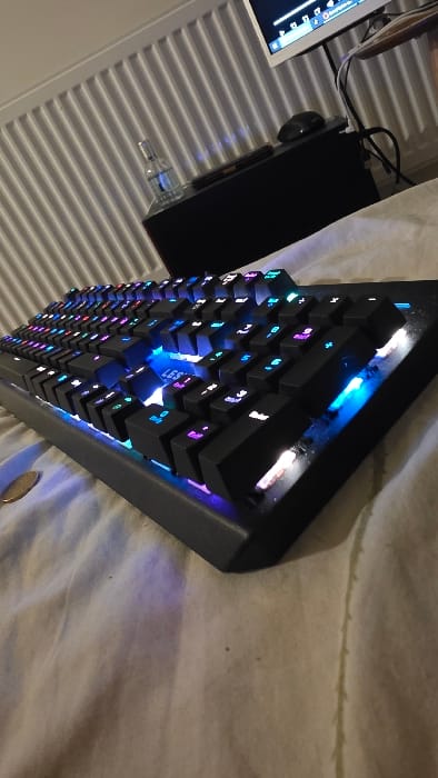 The image depicts an indoor scene in Heusden, Belgium, focused on a black mechanical keyboard with active multicolor RGB backlighting, positioned on a surface covered with creased, light-colored fabric. The keyboard keys illuminate in various hues, including prominent blues, purples, and cyans, with intense blue light visible from beneath the spacebar and right shift keys. A partially visible, round, metallic object is situated on the fabric to the left of the keyboard. In the midground, a dark, rectangular electronic unit supports a clear glass bottle containing a blue liquid, a dark-colored computer mouse, and a dark remote control. Behind these items, a portion of a light-bezel flat-screen monitor displays a graphical user interface, surrounded by multiple dark cables. A white, vertical-panel radiator is mounted on a light-colored wall in the upper left background. No individuals are present in the scene.