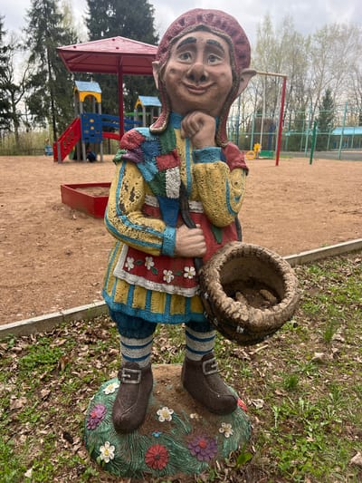 The image shows a garden gnome statue in the city of Sanatoriya Yumatovo Imeni 15-Letiya Bassr, Russia. The gnome is wearing a patchwork jacket of yellow, red, and blue, with striped blue and white pants and brown boots. It has a red hat and is holding a dirty, empty basket. The gnome is standing on a base decorated with flowers. The background shows a playground with various structures, and trees. The ground is covered with sand and some vegetation.