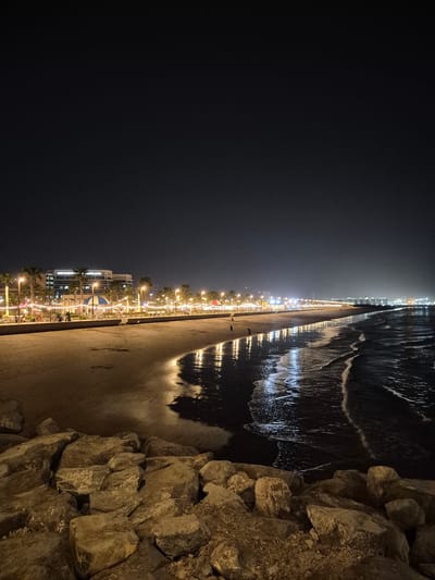 An aerial view captures the Fujairah Corniche at night. The scene is dominated by the dark sky, transitioning into a dimly lit cityscape along the coastline. A sandy beach stretches across the foreground, with the ocean waves gently lapping at the shore, reflecting the numerous lights from the promenade. The promenade is lined with palm trees and illuminated by strings of fairy lights and streetlamps. Several small tents or kiosks are visible along the promenade, suggesting a lively atmosphere. A modern, multi-story building stands prominently in the background, its windows glowing. A few individuals can be seen walking or standing on the promenade and the beach. The foreground is framed by large, rough rocks, possibly part of a breakwater or embankment.