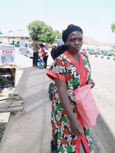 The image depicts an outdoor street scene in Communauté rurale de Payar, Senegal, under bright daylight. In the foreground, a dark-skinned female, approximately 30-40 years old, stands facing the viewer with a neutral expression. She wears a black headwrap and a long, black dress adorned with red and green floral patterns, featuring an orange V-neck and vertical trim. A light pink fabric bag is held in her left hand, resting against her side.
To the immediate left of the central figure, a white sign reads "SOLAKEM COMMUNICATIONS POS AVAILABLE HERE," affixed to a crude wooden stand situated above visible refuse. The paved street extends from the midground into the background. A silver minivan is parked on the street in the mid-left area. Two additional individuals are visible on the sidewalk near the minivan: one wears a brown patterned top and dark trousers, while the other, possibly a child, wears a red and white top and holds a blue bag.
In the background, a line of approximately five light-green, three-wheeled passenger vehicles (tuk-tuks) is parked along the right side of the road, adjacent to a low, light-colored building or wall. Green trees and additional buildings are visible in the distant background, all under a clear sky.