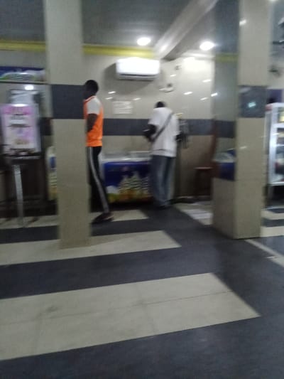 The image shows the interior of a store in Ovwian, Nigeria, located at latitude 5.503 and longitude 5.799. The store has a tiled floor with alternating black and white stripes, and there are pillars on either side of the aisle. Two men are visible, one wearing an orange shirt and black pants and the other wearing a white shirt and grey pants. They are both blurred and moving around, seemingly engaged in a transaction or conversation.  A large blue cooler can be seen behind the men, with some items inside. The room is brightly lit with a white ceiling. The men's actions are unclear due to motion blur. There are several shelves on the left side of the room, though the items on them are not discernible. The overall scene suggests a busy and somewhat chaotic environment.