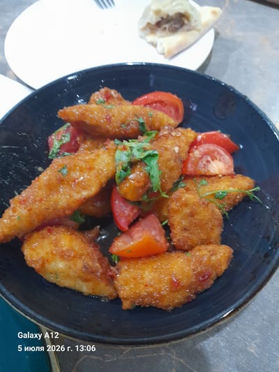 A close-up, overhead shot showcases a dark blue bowl filled with golden-brown, breaded chicken strips. The chicken pieces are coated in a glistening sauce and interspersed with bright red tomato wedges and chopped green herbs, likely parsley or cilantro. In the upper right portion of the image, a white plate holds a piece of flatbread that has been torn open, revealing a meaty filling. The background is a textured surface, possibly a tabletop, with streaks of gray and brown. In the bottom left corner, superimposed text reads "Galaxy A12" and "5 июля 2026 г. 13:06". The scene appears to be indoors, likely in a restaurant or home setting in Kara-Köl City, Kyrgyzstan. There are no people or animals visible in the image.