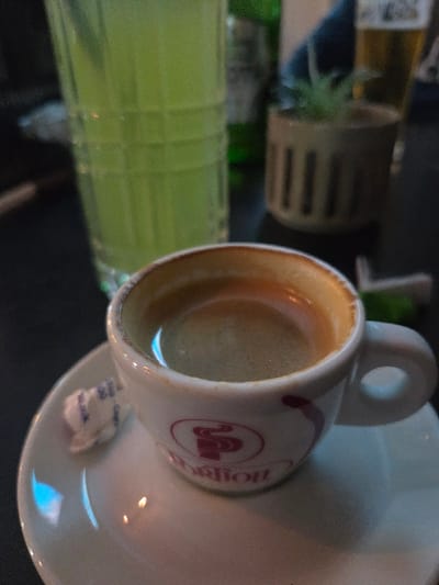 The scene depicts a tabletop arrangement in Negorci, North Macedonia. In the foreground, a white ceramic espresso cup, bearing a red stylized 'S' logo and the text "Pontion", contains dark coffee topped with crema. The cup rests on a matching white ceramic saucer, which also holds a small, white, rectangular sugar packet. Immediately to the left of the espresso cup, a tall, clear glass contains a bright green liquid. The background is out of focus, revealing a small, brown, slatted cylindrical pot holding a green succulent plant. Further obscured elements include a green bottle and a glass containing an amber liquid, consistent with beer. No individuals are visible. The surface supporting these items is dark, suggesting a table or bar top, under what appears to be ambient, somewhat dim lighting.