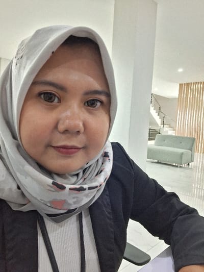 A woman wearing a light gray hijab poses indoors. The hijab has a subtle, patterned design. She is facing the camera with a slight smile. She is wearing a black jacket over a white shirt with horizontal black stripes. In the background, a modern staircase and a light gray sofa are visible. The room appears to have white walls and good lighting.