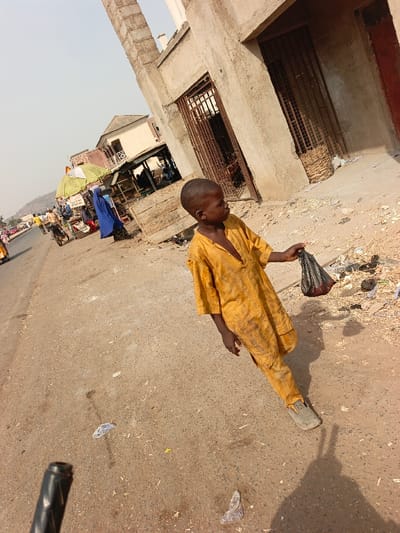 A young boy wearing a mustard-yellow, traditional robe and pants is walking on a dirt and paved street. He holds a small plastic bag. The street is cluttered with trash. In the background, there are several market stalls covered by large umbrellas and a few buildings. A person in a blue robe stands by one of the stalls. The sky is overcast and bright.