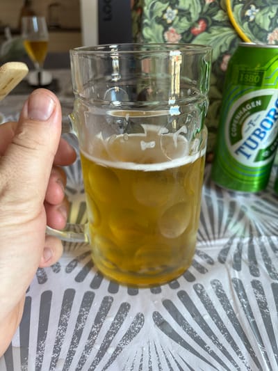 An outdoor close-up shot shows a person's hand holding a beer mug filled with golden lager. The mug is clear glass with a handle and is about two-thirds full. Bubbles are visible on the surface of the beer. Behind the mug, a green can of Tuborg beer is partially visible. In the background, out of focus, is a table with a patterned cloth, and beyond that, a blurred glimpse of a wine glass with a yellowish liquid. The lighting suggests a daytime scene. The image does not provide any information that can place it in the city of Cork, Ireland.