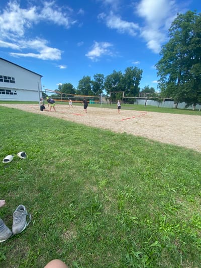 The image shows a sandy volleyball court with a net, set up in a grassy area under a bright blue sky with scattered white clouds. There are trees in the background and a white building with windows on the left. Several people are playing volleyball on the court. On the right side of the image, there are two pairs of shoes on the grass, and a person's bare feet and legs are visible. The scene appears to be during daylight on a pleasant day, likely outdoors in a recreational area. The overall mood seems casual and active, with people enjoying a game of volleyball. No visible text is present in the image.