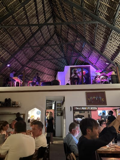 This image captures the interior of a vibrant restaurant or bar, likely in Michamvi, Tanzania, during the evening. The scene is dominated by a distinctive, high-pitched thatched roof (makuti), constructed from interwoven straw or palm fronds supported by dark wooden beams.
On the left side of the image (when oriented correctly), a performance area or stage is visible, illuminated by purple ambient lighting. Here, a drum kit with metallic cymbals and purple-toned drums is set up. A large, colorful painting, possibly depicting a figure or scene, adorns the wall behind the stage, also bathed in purple light. Below this, sound equipment or speakers are visible.
To the right, a white wall separates the stage area from the main seating area. This wall features framed artwork, including a piece with stylized figures and smaller pictures arranged within an arched alcove. Several mirrors on this wall reflect parts of the room and patrons.
Numerous people, guests of the establishment, are seated at tables in the foreground and mid-ground. They appear to be socializing and enjoying their evening. Notable individuals include a man with dark hair in a dark shirt, another man in a light blue shirt, and various other patrons engaged in conversation. A woman with blonde hair is visible as a reflection in one of the mirrors. The presence of bottles and glasses suggests a bar or dining setting.
The overall atmosphere is lively and relaxed, indicative of an evening event, possibly featuring live music, given the stage setup and dynamic lighting. No discernible text is visible in the image.