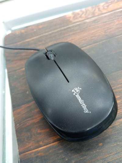 A black, wired computer mouse rests on a wooden surface. The mouse has a textured grip and a scroll wheel. A white logo with a cartoon character and the text "smartbuy" is visible on the surface of the mouse. The wooden surface appears to be a tabletop or desk, with visible grain patterns. The background is blurred, suggesting a shallow depth of field. No people are present in the image. The location is not discernible from the image itself, and there is no information to connect it to the city of Kuybyshev, Russia.