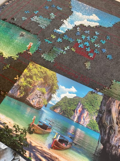 A partially assembled jigsaw puzzle lays on a grey puzzle mat with red grid lines. The puzzle appears to depict a tropical beach scene with boats, cliffs, lush vegetation, and a blue sky with clouds. Loose puzzle pieces in varying shades of blue, green, and brown are scattered around the partially assembled areas. The puzzle is not located in Bukulti, Latvia.