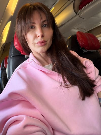 A young adult female is depicted in a close-up, chest-up shot, presumably a selfie, inside an airplane cabin in Bol'shie Kabany, Russia. She has long, dark brown hair styled with bangs, framing her face, and her hair falls over her shoulders and chest. Her eyes are light-colored, and she maintains a direct, neutral gaze at the camera. Her lips are full and slightly parted. She is wearing a light pink, long-sleeved quarter-zip sweatshirt, with a white undershirt visible at the neckline.
The background reveals the interior of a commercial aircraft. To her immediate left, multiple aircraft windows are visible, through which bright, diffused light enters the cabin. Behind her is a black airplane seat with a prominent, integrated red leather headrest. Another similar seat with a red headrest is partially visible further back and to the right. In the upper right quadrant of the image, standard white overhead luggage bins are visible, with a red bag or object partially stored within one. The number "50" is clearly marked on the underside of an overhead bin. Faintly, the profile of another passenger can be seen in the distant background beyond the rightmost seat. The cabin lighting appears warm and diffused.