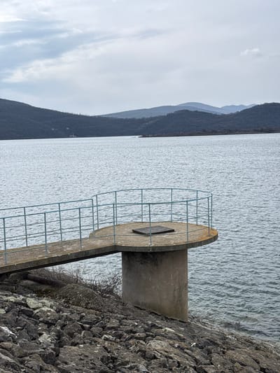 The image depicts a tranquil outdoor scene on an overcast day, located near the city of Баня, Bulgaria, featuring a concrete observation platform extending over a large body of water, likely a reservoir or dam.
In the foreground, a steeply sloped embankment covered with dark, irregularly shaped rocks and minimal dry vegetation transitions into the water. A concrete walkway, supported by a robust, cylindrical concrete pillar that disappears towards the water level, extends from the embankment to a circular concrete platform. Both the walkway and the platform are equipped with a light blue metal railing, consisting of multiple horizontal bars and vertical supports, encircling the platform and lining the walkway. A dark, square metal hatch is visibly embedded in the surface of the circular platform.
The midground is dominated by a vast expanse of grey-blue water, exhibiting subtle ripples across its surface. In the background, a series of dark blue-grey hills and mountains with indistinct tree cover rise, defining the distant shoreline. A faint, light-colored structure, possibly a building, is discernible on one of the hillsides to the left. The sky is uniformly overcast, presenting a soft, diffused light across the entire scene. No people or animals are visible.