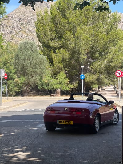 In Port De Pollença, Spain, a maroon convertible Alfa Romeo Spider, license plate M44 RSD, is positioned at a junction. The car faces away from the viewer, angled towards the right, with its top down. A person with blonde hair is visible in the driver's seat, wearing a green top, and looking forward.

The scene unfolds on a paved road with a pedestrian walkway to the right, bordered by trees and a low wall. A mountain range with rocky terrain forms the backdrop. Traffic signs are present: a blue circular sign with a white right arrow, and a red circular sign with a white horizontal bar and a red diagonal bar indicating no entry or prohibition of a turn. The foreground is partially shaded by dappled sunlight filtering through foliage, suggesting a sunny day. The road shows signs of wear, with cracks visible in the asphalt.