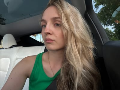 A young Caucasian woman with long, wavy blonde hair is shown from the chest up, seated in the passenger seat of a vehicle. She is wearing a green tank top and a rose gold necklace with a small pendant. The interior of the car is visible, including white leather seats and a dark-colored dashboard and pillar. The car has a panoramic glass roof, through which the sky and trees are visible. The scene appears to be in a suburban or residential area, with lush green trees and a cloudy sky suggesting an outdoor setting. The woman is looking slightly away from the camera with a neutral expression.