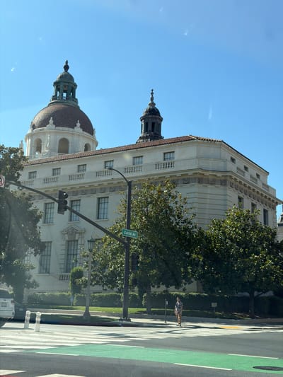 The image shows the exterior of the Pasadena City Hall, a prominent building in Pasadena, California, United States of America. The building features a large, red-tiled dome with a smaller, dark-colored dome on top, as well as a long, cream-colored facade with multiple windows and a decorative balustrade. A street lamp and traffic signals are visible in front of the building, along with a street sign indicating "Euclid Av." Trees with green foliage are planted in front of the building. In the foreground, a crosswalk with green accents is painted on the asphalt. A person is walking across the crosswalk. A white SUV is parked on the left side of the image. The sky is clear and blue.