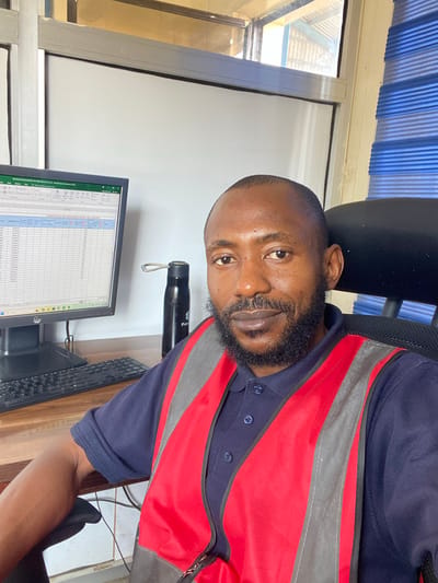 A man sits at a desk in an office setting, facing the camera. He has dark skin, a beard, and is wearing a navy blue polo shirt with a red and gray safety vest over it. The man's head and upper torso are visible. He appears to be looking directly at the camera with a neutral expression. To his left, on the desk, is a computer monitor displaying a spreadsheet. A black water bottle with a tan cord is standing upright next to the monitor. A black keyboard is also visible on the desk. The background shows a window with blue blinds, and part of an office chair. The scene is indoors, and no specific time of day or weather conditions are discernible. The location context is Kinkinau, Nigeria. Visible text on the computer monitor includes "TECHNOLOGICAL REPORT FOR NNN.EU 21/22 (31/01/22) - Heat Product Application Data".