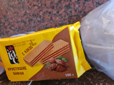 An outdoor, eye-level, close-up shot captures a package of "Tac" brand wafers with cocoa flavor. The packaging is predominantly yellow and brown, with a brown design featuring cocoa pods, beans, and powder. Text on the packaging includes "PRO. 14.07.2026," "EXP. 14.07.2027," "с Какао" (with cocoa), "ХРУСТЯЩИЕ ВАФЛИ" (crispy wafers), and "180 г" (180g). The background is out of focus, showing a speckled reddish-brown surface, possibly a granite countertop, and a white plastic bag on the right side. The scene appears to be set in Saray, Azerbaijan, based on the provided context.