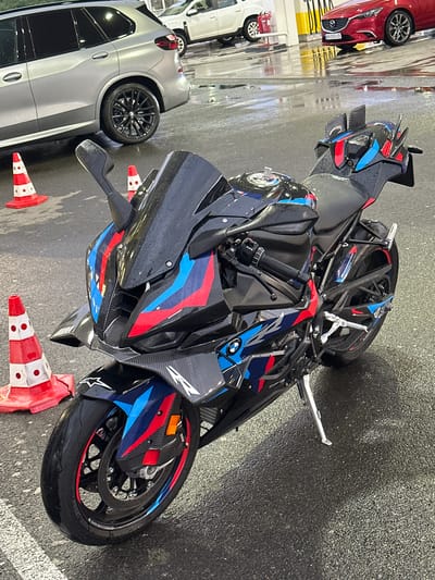 This image depicts a black BMW sport motorcycle with a distinctive red, blue, and white graphic design parked on wet asphalt. The motorcycle is positioned in the foreground, angled towards the right side of the frame. It features carbon fiber accents, a raised rear section with small winglets, and a low-slung front fairing with dual headlights. In the background, several cars are visible, including a silver SUV, a white sedan, and a red Mazda. Orange and white traffic cones are placed around the motorcycle. The environment appears to be an outdoor parking area or lot, possibly at a gas station or car wash, given the wet surface and the presence of vehicles that suggest a public thoroughfare or commercial establishment. The lighting suggests daylight, with reflections visible on the wet pavement. The scene is set in Drazhevo, Bulgaria.