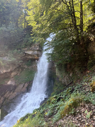 The image depicts a natural waterfall cascading down a rocky cliff face, surrounded by lush green foliage and trees. The scene is illuminated by sunlight filtering through the leaves, creating dappled light and shadows. Several individuals are visible near the waterfall's edge, observing the spectacle. One person in a red shirt is standing on a wooden walkway to the right of the main water flow. On the left side, two individuals, one in white and a smaller figure, stand on a rocky ledge. Wooden railings and a metal fence are present along the pathway. The environment suggests a natural park or a hiking trail in a mountainous region. The location is identified as the Giessbach Falls in Axalp, Switzerland.