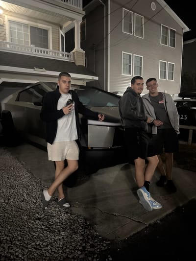 Three young adult males stand next to a silver Tesla Cybertruck parked on a gravel driveway in front of a two-story residential building at night in Ocean City, USA. The man on the left is wearing a black zip-up jacket over a white t-shirt and light-colored shorts. He is holding a smartphone in his right hand and looking at the camera. The man in the middle is wearing a dark grey hoodie and black shorts, with his right arm resting on the Cybertruck. The man on the right is wearing a grey zip-up hoodie over a dark grey t-shirt and black shorts, with his left arm resting on the Cybertruck. The background shows the facade of a multi-story house with multiple windows and a dark sky. The Cybertruck is prominently positioned in the foreground, partially obscuring the vehicle's wheels and the driveway. The overall scene suggests a casual, outdoor gathering.