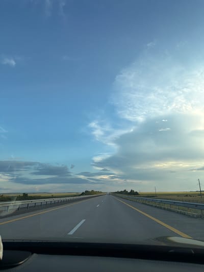 The image depicts a highway scene, likely taken from the perspective of a vehicle driving on the road. The highway stretches into the distance, with clear lane markings and a guardrail on the right side. The surrounding landscape appears to be flat and rural, with fields of grass or crops on either side of the road. Power lines are visible in the distance. The sky is partly cloudy, with a mix of blue sky and white and gray clouds. The lighting suggests it might be late afternoon or early evening. The location is specified as the city of Minovka, Kazakhstan. There are no people or specific subjects or objects that stand out besides the road and the landscape. The image is an objective record of a road trip scene.