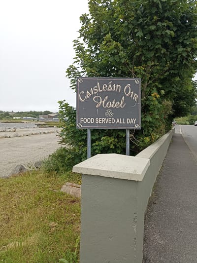 The image shows a sign for "Caisleán Óir Hotel" with "FOOD SERVED ALL DAY" below it. The sign is dark with light-colored lettering and decorative elements, including what appears to be a Celtic knot. It is mounted on two metal poles and partially obscured by green foliage from a tree. In the foreground, there is a low concrete wall painted a muted green, topped with a lighter colored coping. To the left of the sign, a gravel parking area is visible, bordered by a line of stones. Beyond the parking area, there are some buildings and greenery, suggesting an outdoor setting. To the right of the wall, an asphalt road is visible, with a few cars in the distance. The sky is overcast. The location is Annagry West, Ireland.
