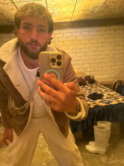 A male individual is depicted in a mirror selfie taken indoors in a room in Uyuni, Bolivia. He has light brown hair, a beard, and a nose ring on his left nostril. He wears a brown jacket with a cream-colored sherpa lining, a white long-sleeved shirt, and light-colored pants. A beaded bracelet is visible on his right wrist as he holds a smartphone with a gold-colored case and a circular, multi-colored pop socket/grip to capture his reflection. The room features a light-colored brick wall in the background and a ceiling composed of decorative, reflective panels with faint patterns. To the right, a bed is present, covered with a dark blue bed skirt and a patterned blue and brown blanket. A pair of white rubber boots stands upright on the floor near the bed. The floor itself is light-colored and highly reflective, particularly around the subject's feet, showing some unevenness or a white residue. The scene is illuminated by warm, artificial light.