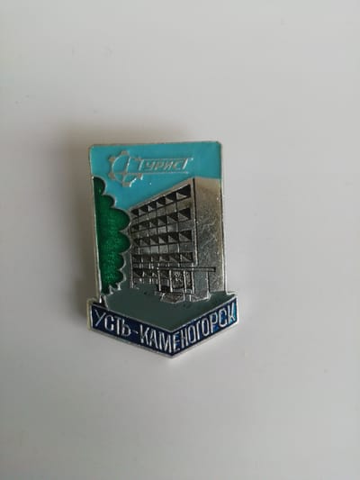 The image shows a lapel pin with a depiction of a building and trees. The top of the pin features a stylized gear and the word "ТУРИСТ" (Tourist) in silver lettering on a blue background. Below this is a silver building with a patterned facade, a green tree on the left side, and a dark blue banner at the bottom with the Cyrillic text "УСТЬ-КАМЕНОГОРСК" (Ust-Kamenogorsk). The pin is made of metal with enamel inlays. The background is a plain, light-colored surface, likely a table or desk. Ust-Kamenogorsk is a city in Kazakhstan, located in the Altai region.