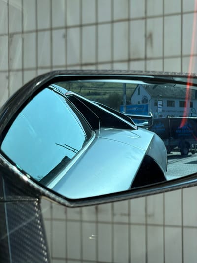 The image displays a close-up view of a vehicle's driver's side rearview mirror, which features a carbon fiber housing on its visible lower-left portion. The mirror reflects the rear-left quarter of a light-colored (silver or light gray) high-performance sports car, characterized by sharp angular bodywork, a slatted engine cover, and a prominent rear spoiler.

Beyond the reflected vehicle, the mirror shows an exterior scene identified as Adenau, Germany. In the background, a green, undulating landscape with a single bare tree is visible under a clear sky. In the mid-ground, a white, two-story house with dark windows and a gabled roof stands, partially obscured by a larger building featuring a prominent blue sign. Mirrored German text, including "VERTRIEB" (distribution/sales), is discernible on this blue sign. To the right, a dark utility trailer, possibly covered with a black tarpaulin, is parked on a paved surface. Mirrored German text, including "SAMMELHANDEL" (collective trade/wholesale), is visible on the trailer's side. The area immediately behind the mirror and forming the upper portion of the main image reveals a light-colored wall with a distinct grid pattern, possibly composed of tiles or panels. No individuals or active interactions are visible in the scene.