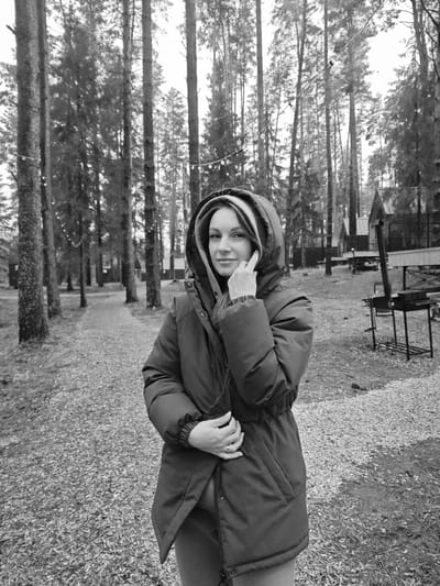 This black and white image captures a young adult woman standing in a forested, outdoor setting, identified as Мишкино, Russia. The woman is positioned centrally, facing slightly to her right but looking directly at the camera with a gentle, calm expression, a subtle smile playing on her lips. She is wearing a thick, dark-colored hooded winter jacket, cinched at the waist, with the hood up around her head. Her right hand is raised to her face, with a finger touching her cheek, while her left hand holds her right arm.
The background features numerous tall, slender conifer trees, indicating a dense forest or wooded area. A path covered with fallen leaves or wood chips extends into the distance between the trees. In the mid-ground and background, several rustic wooden cabins or small houses with dark roofs are visible, suggesting a recreational area or holiday village. To the right, an outdoor grilling station with a barbecue grill or fire pit and an accompanying table or bench is present. String lights are strung between some of the trees, hinting at an inviting evening ambiance.
The lighting is soft and diffused, typical of an overcast day, with no harsh shadows. The woman's attire and the ground cover of fallen leaves suggest a cool season, likely autumn or early winter. The scene evokes a sense of tranquility and a connection with nature within a recreational context. No visible text is present in the image.